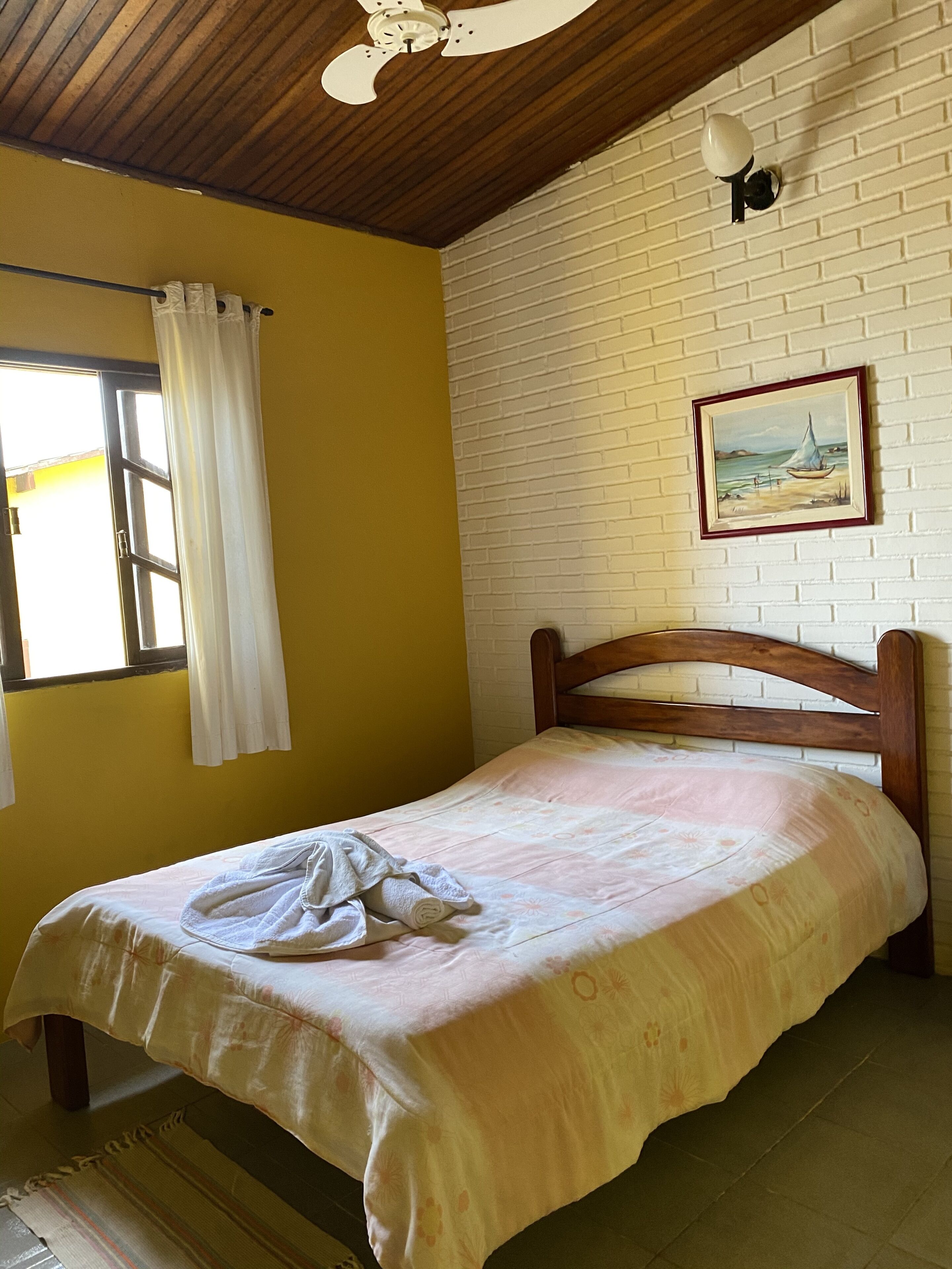 Un lit simple avec une tête de lit en bois, dans une pièce aux murs jaunes et à un mur en briques blanches.