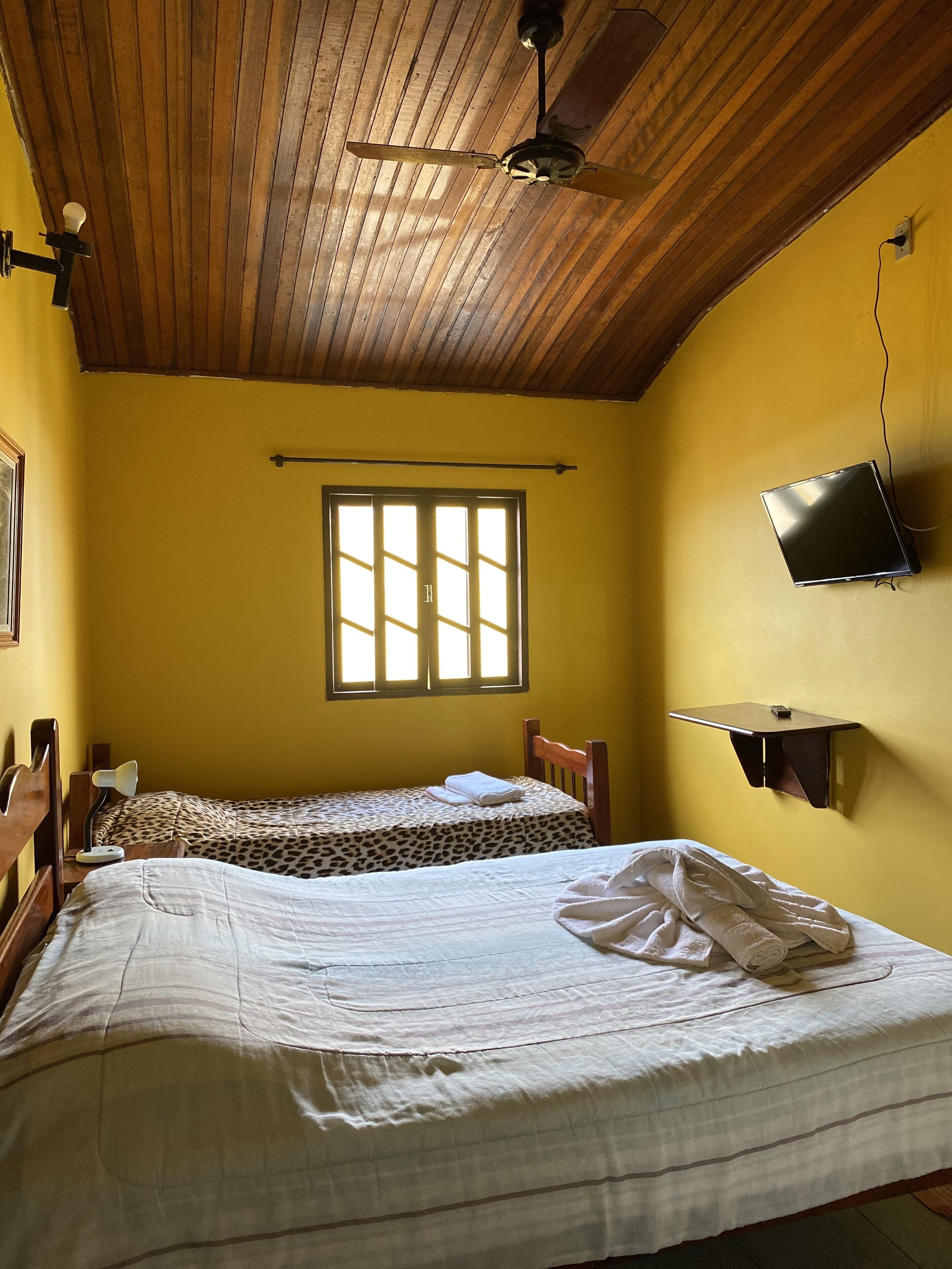 A room with two beds, a ceiling fan, a TV mounted on the wall, and a window with blinds.