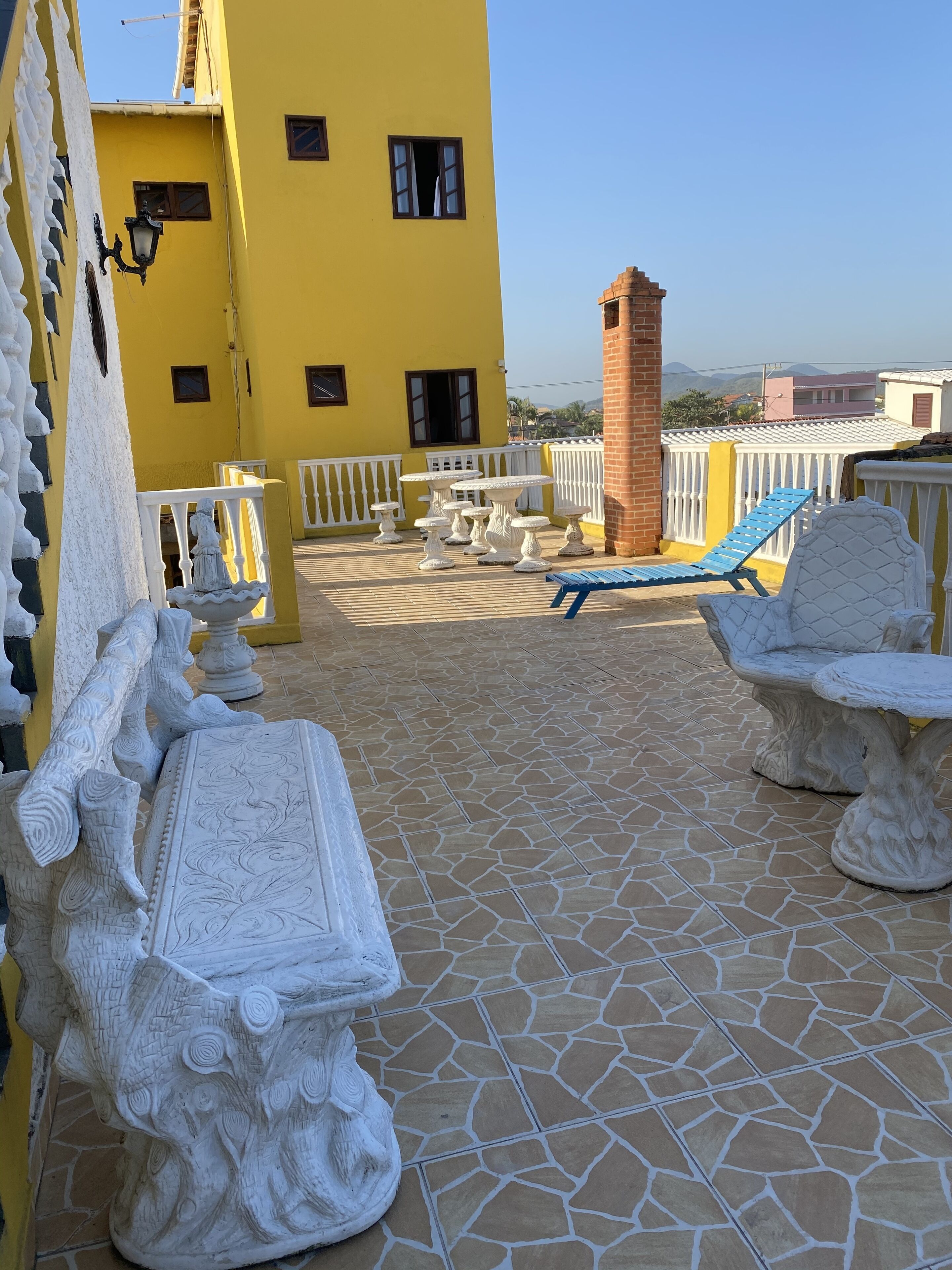 Un bâtiment jaune doté d’un patio carrelé, agrémenté de mobilier en pierre blanche, notamment une table et des chaises, ainsi qu’un fauteuil de jardin bleu.