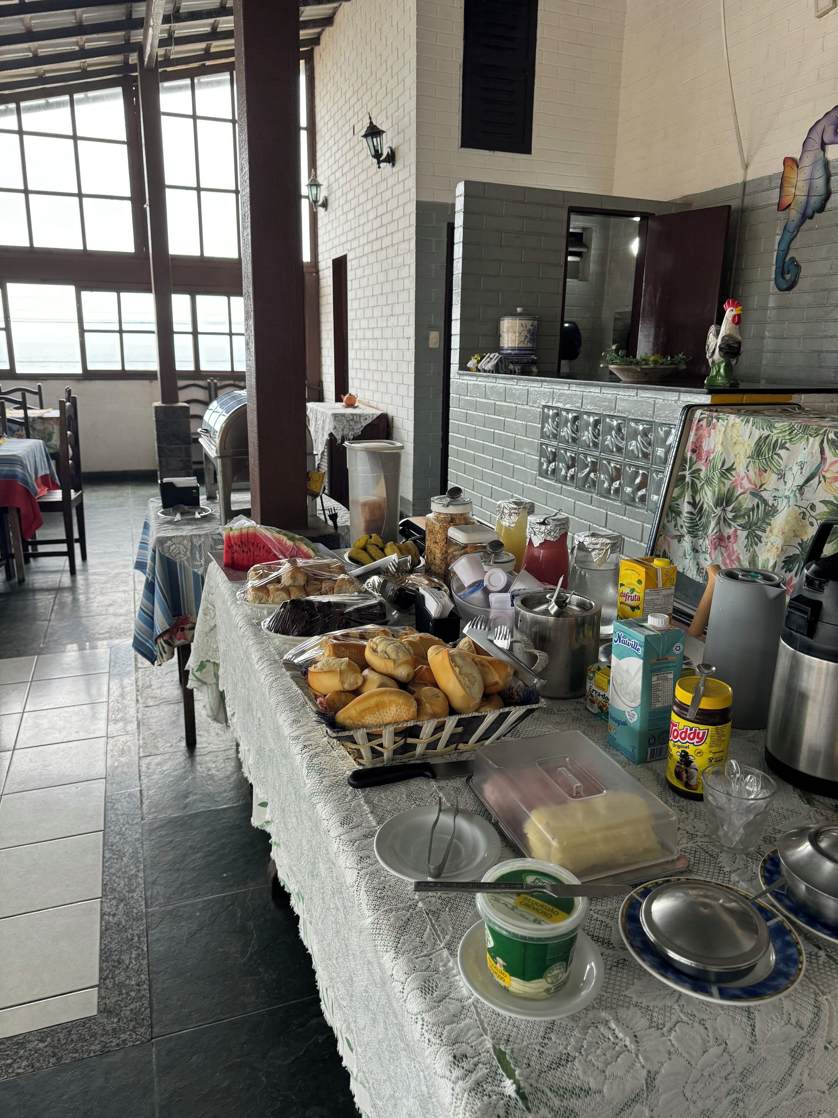 A buffet setup with various food items and condiments on a table, a pizza oven, and a kitchen area in the background.