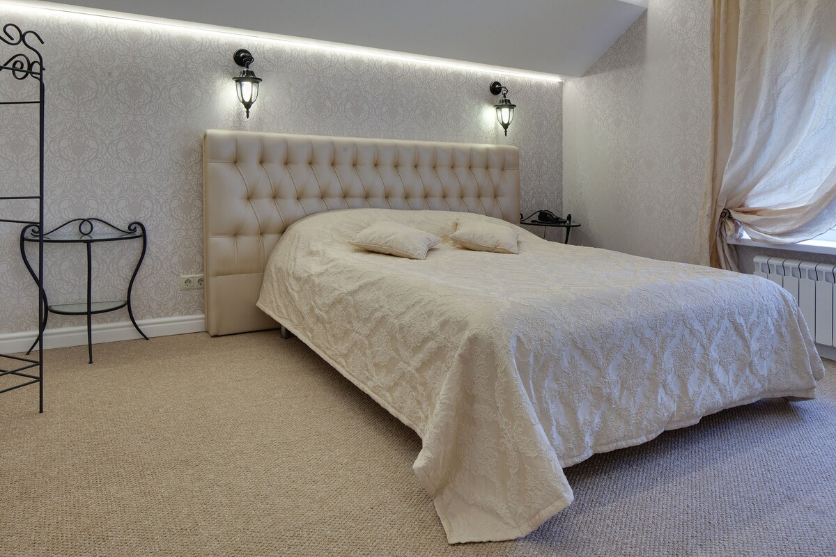 A bedroom with a tufted headboard, two bedside lamps, a bed with a white bedspread, a small bedside table, and a radiator.