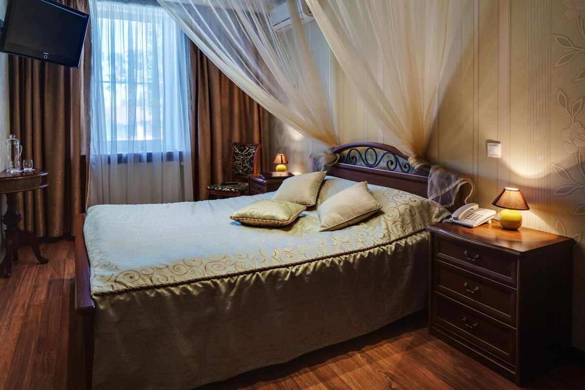 A hotel room with a large bed, a bedside table with a lamp, a TV mounted on the wall, and a window with curtains.