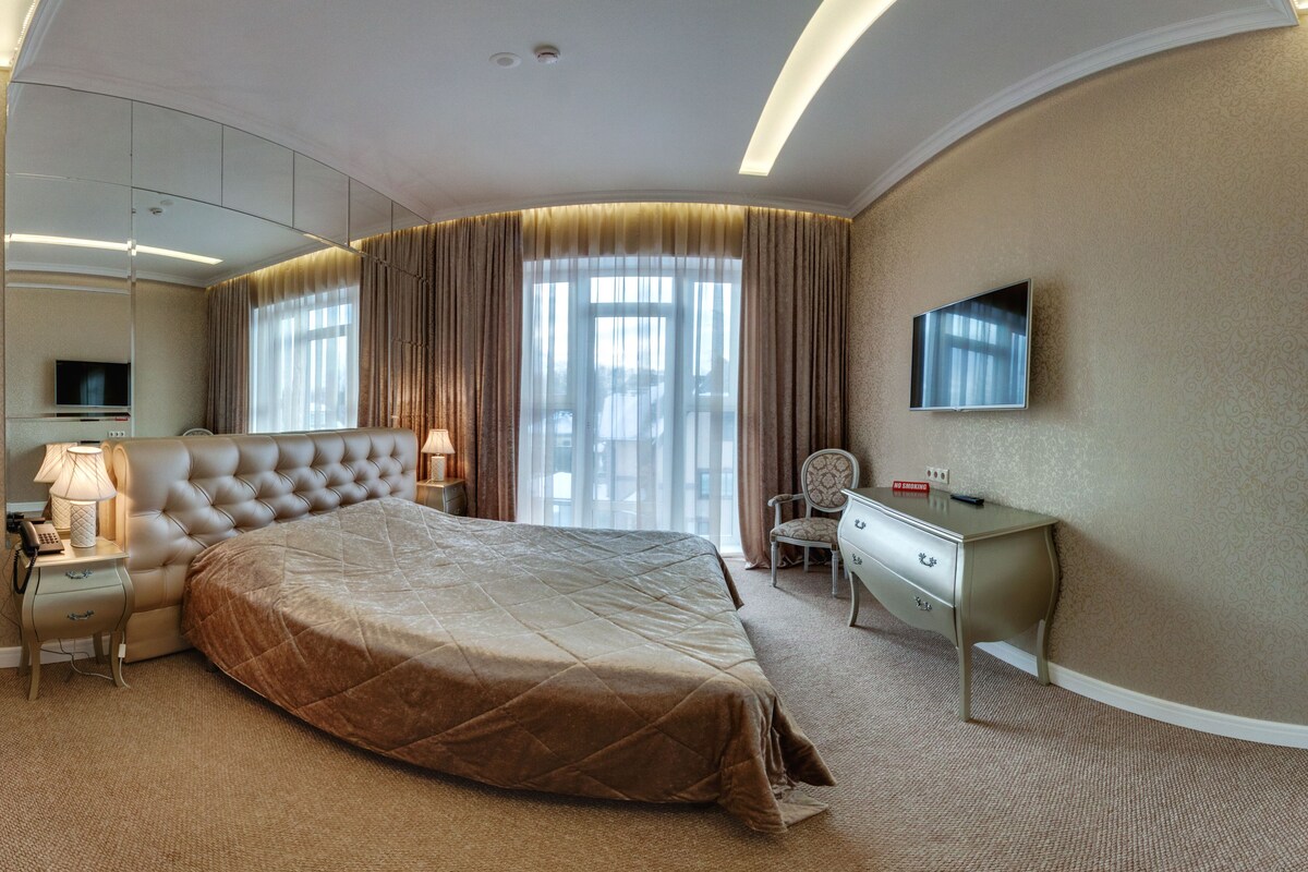A hotel room with a large bed, a desk, a chair, a TV, and a window with curtains.