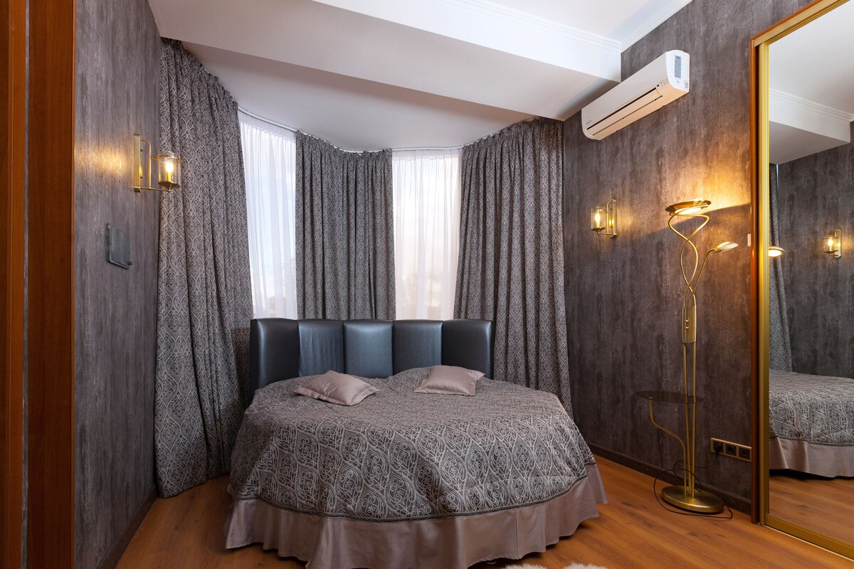 A bedroom with a large bed, dark curtains, a floor lamp, and a mirror.