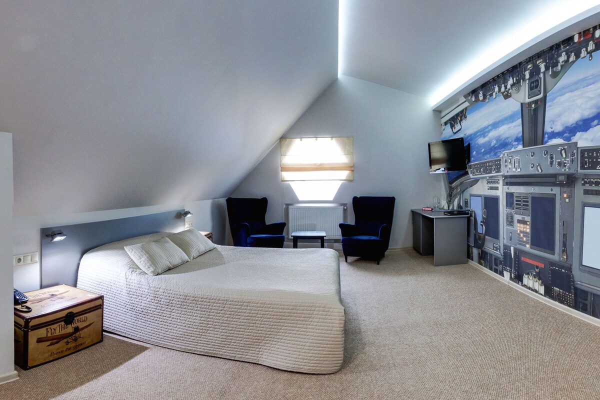 A bedroom with a large bed, a desk, two chairs, a television, and a wall mural.