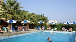 Une piscine entourée de chaises longues et de parasols, avec des palmiers tout autour et un bâtiment en arrière-plan.