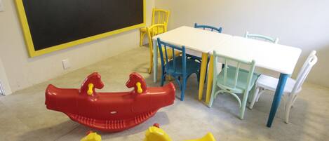 Una habitación con una mesa blanca, sillas azules y amarillas, y un caballo balancín rojo y amarillo.