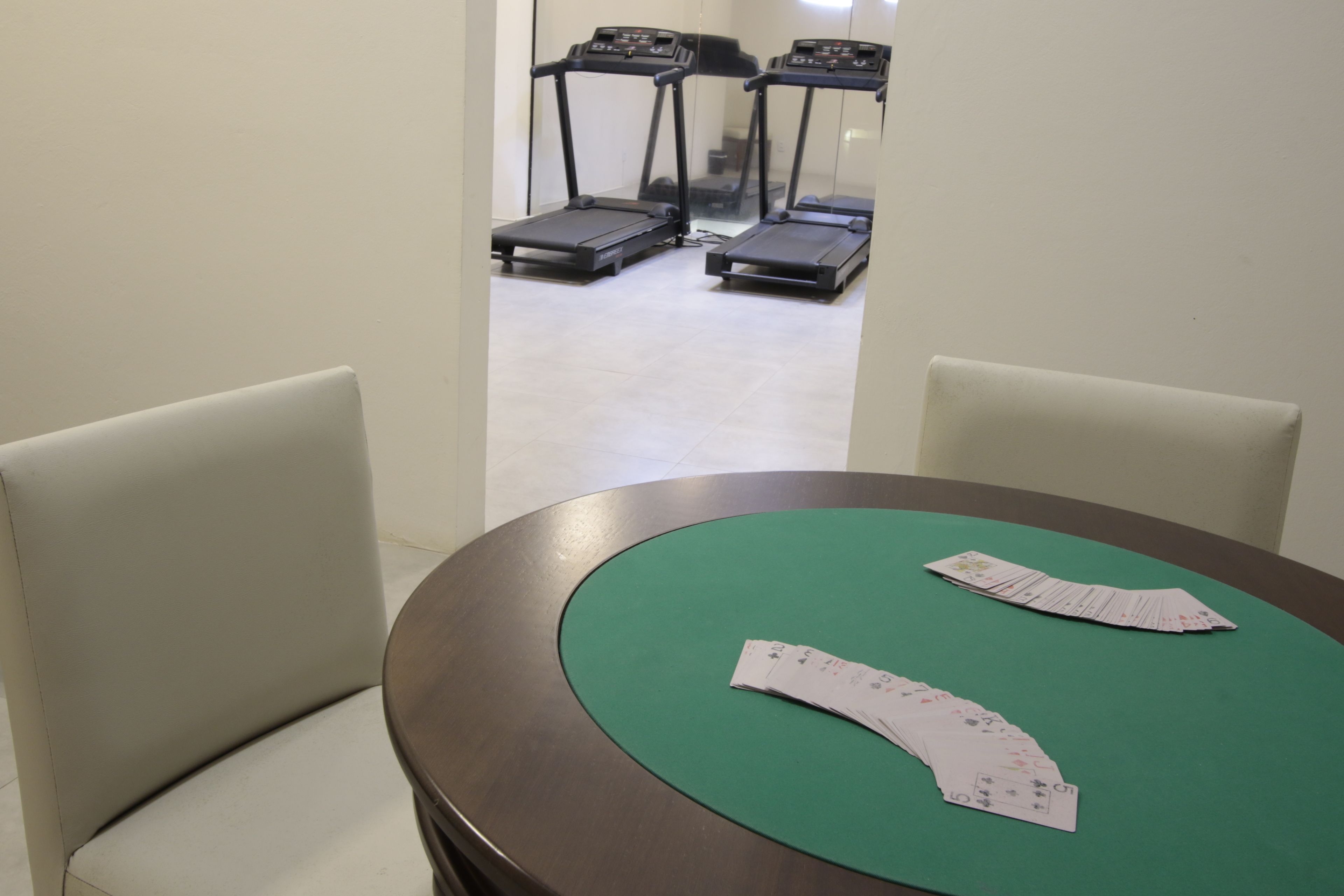 A room with a round table, two chairs, and a green tablecloth. There are three treadmills in the background.