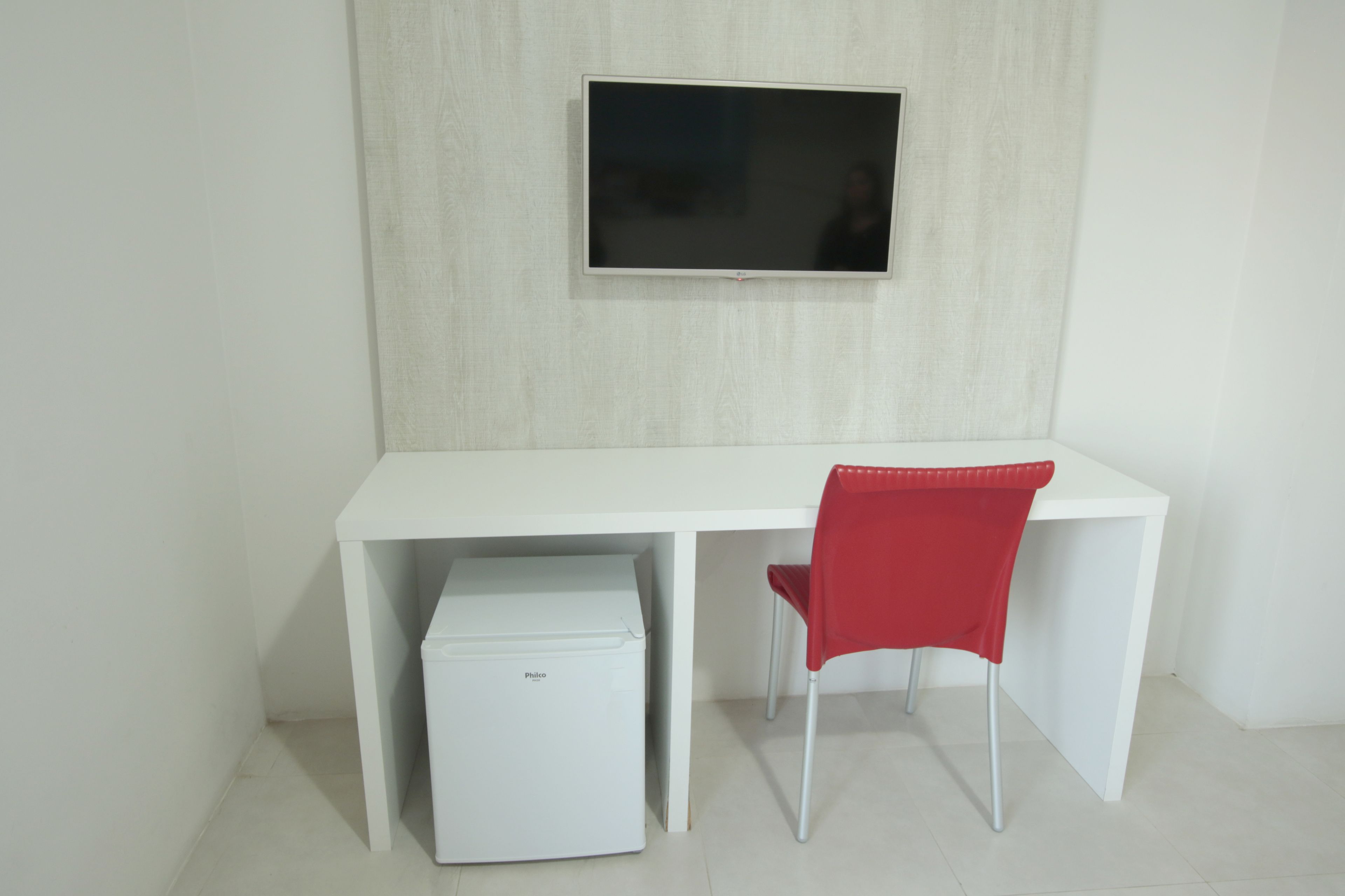 A white desk with a flat-screen TV mounted above it, a red chair, and a white mini-fridge under the desk.