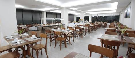 A spacious dining area with multiple tables and chairs, a central serving station, and a large window providing natural light.