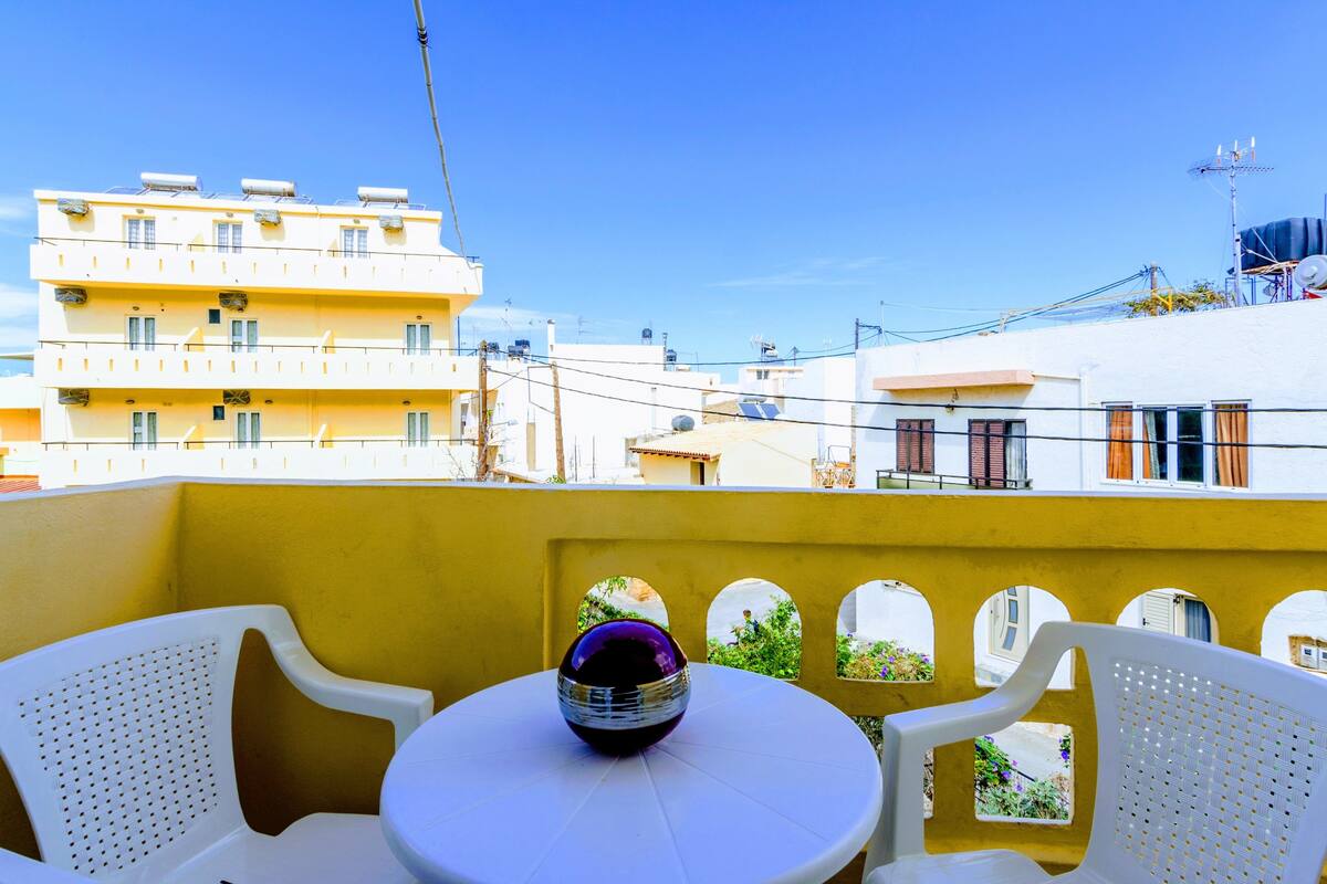Uma varanda com cadeiras e mesa de plástico branco, com vista para um edifício amarelo com várias janelas e varandas.