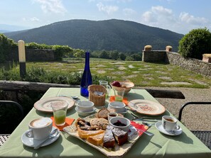 Une table dressée à l’extérieur, offrant une vue sur les montagnes, avec un panier de viennoiseries, des tasses de café et une bouteille d’eau.