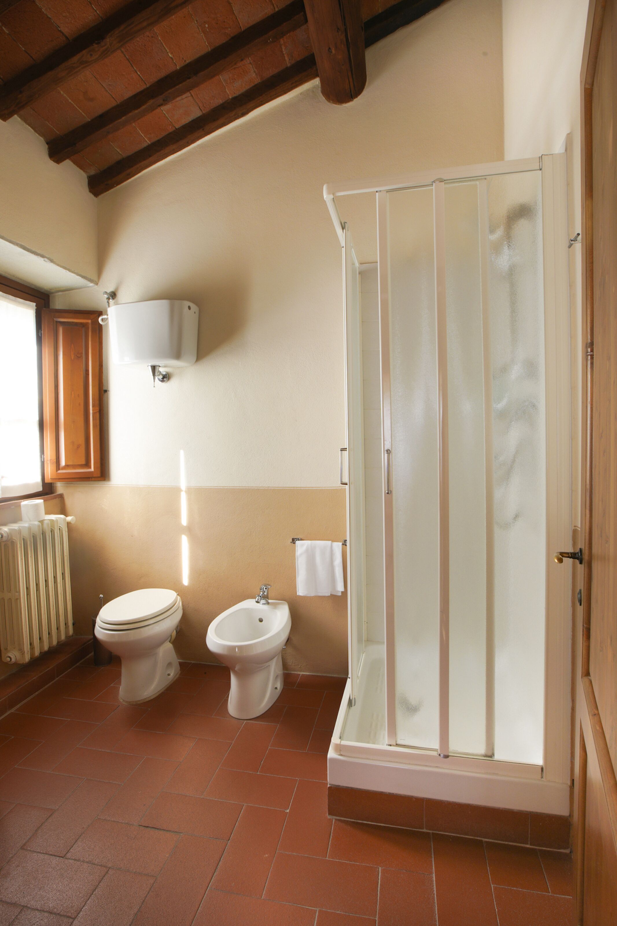 A bathroom with two toilets, a bidet, a glass-enclosed shower, a radiator, and a wooden ceiling.