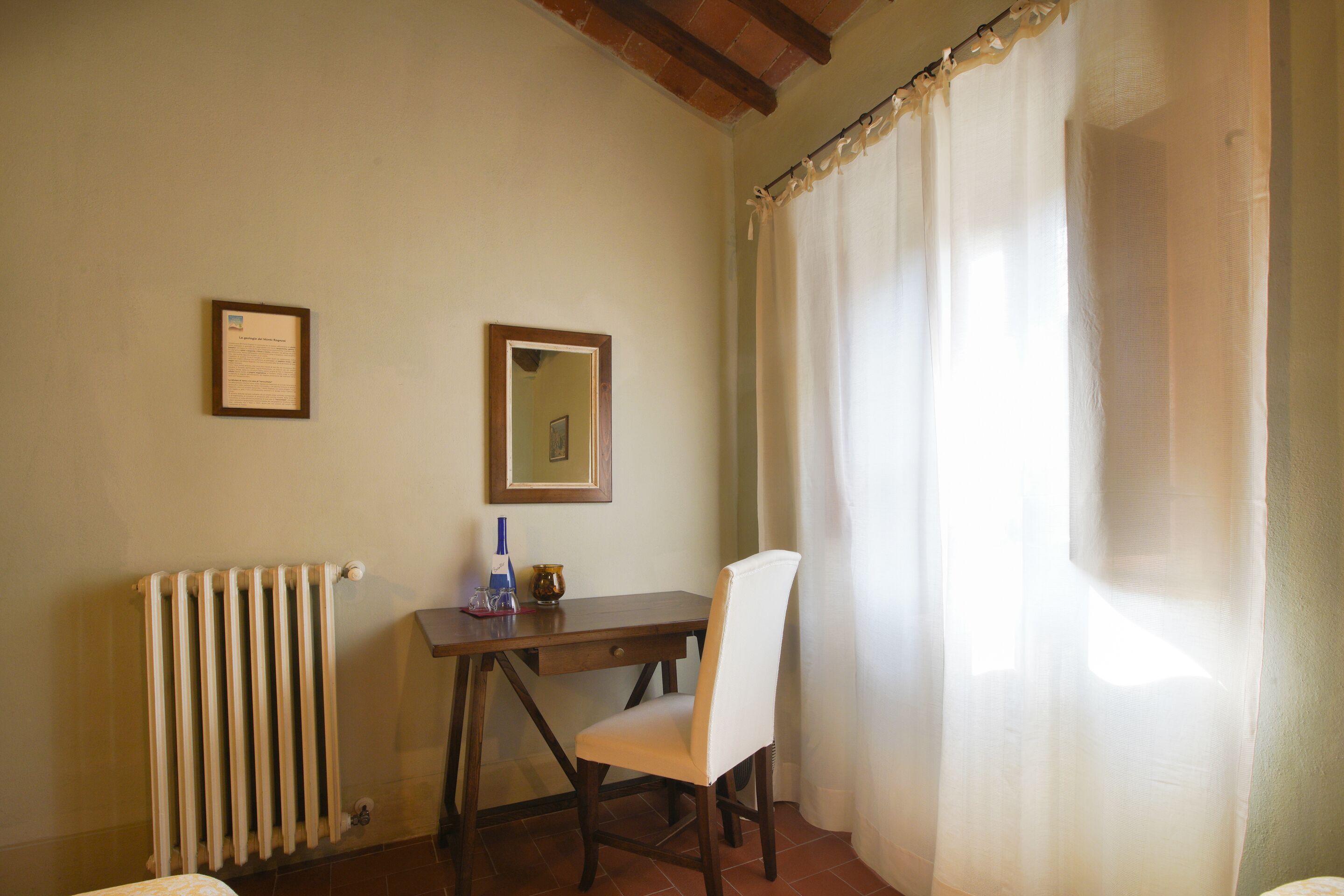 A small room with a wooden desk, a chair, a radiator, and a window with sheer curtains.