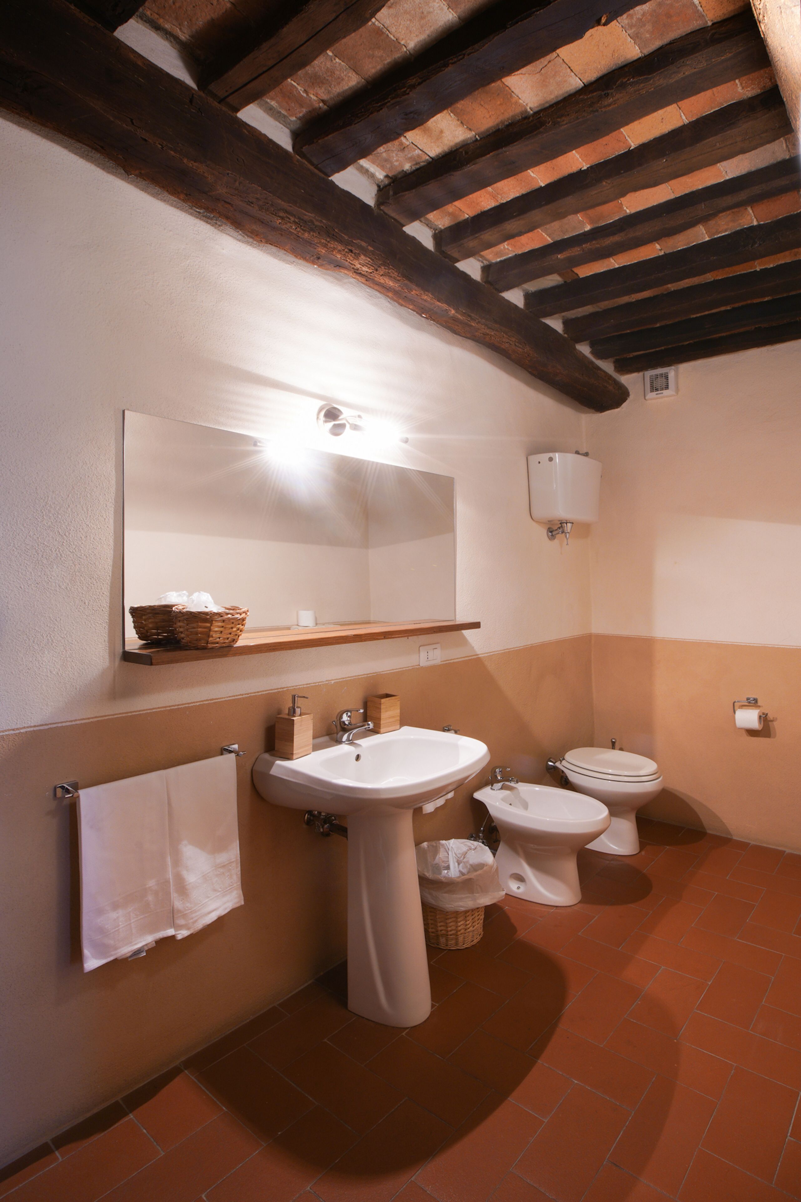 A bathroom with a wooden ceiling, a large mirror, a sink, a toilet, and a shelf with a basket.