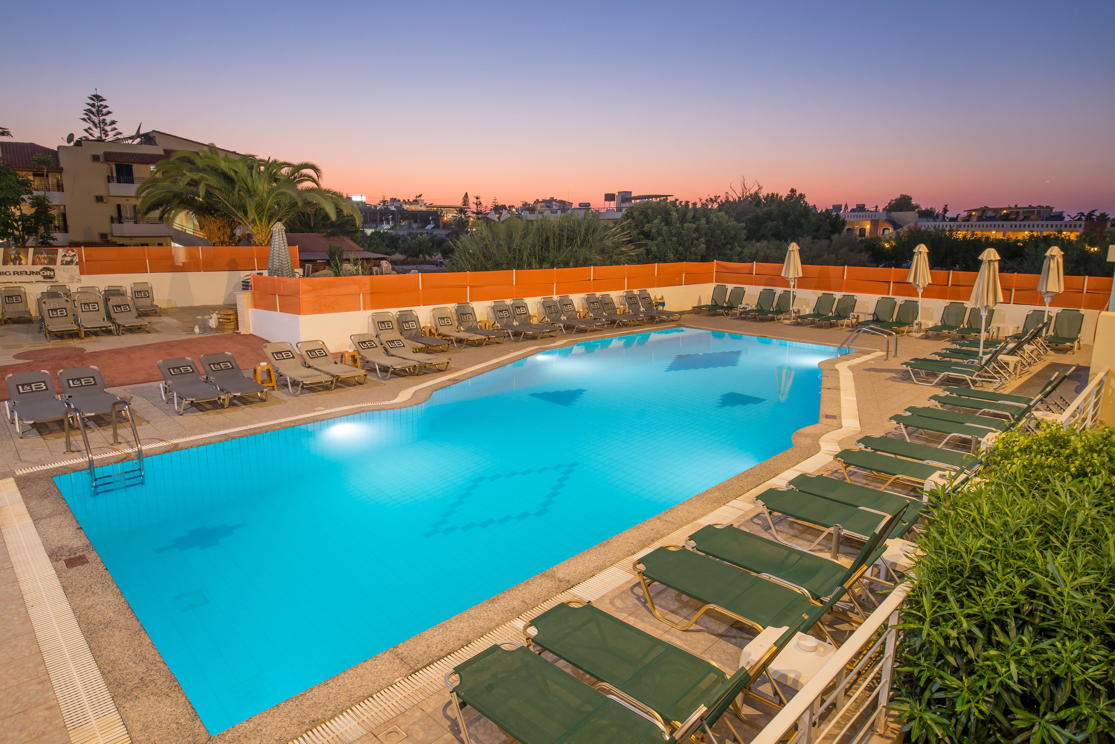 A swimming pool with lounge chairs and umbrellas, surrounded by a fence and palm trees.