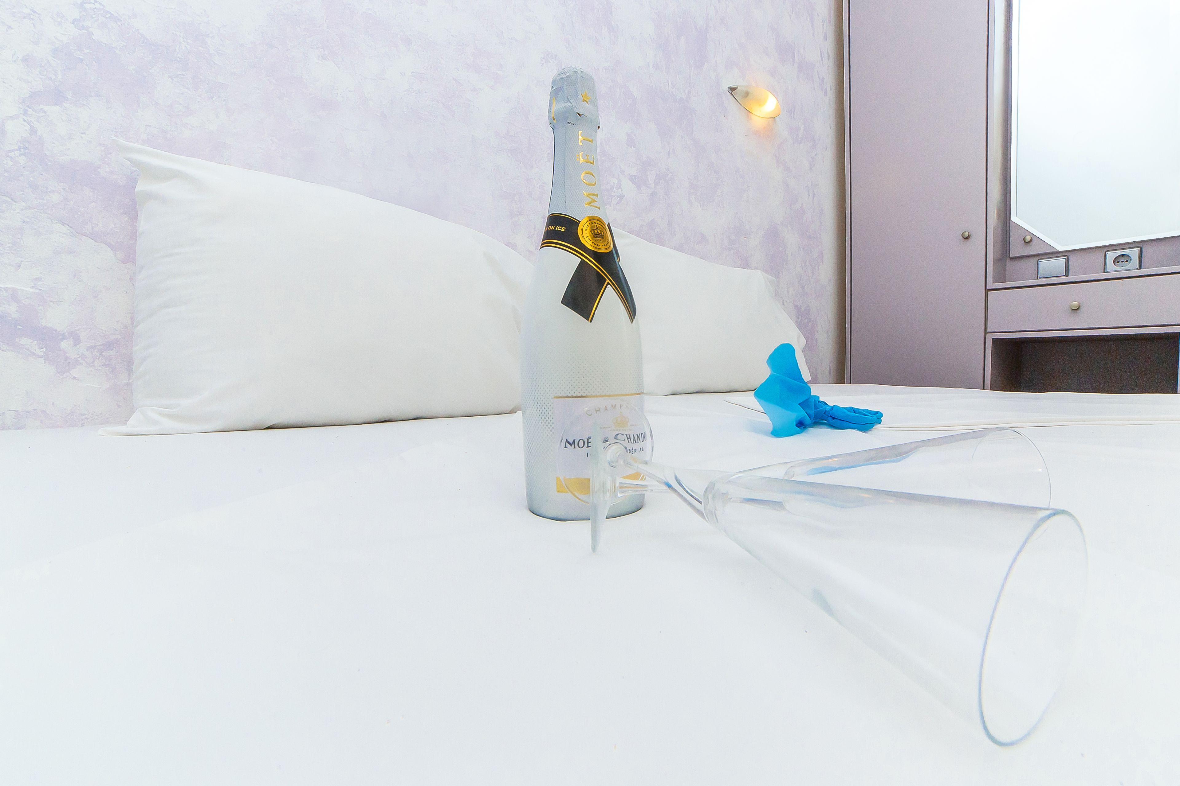 A bottle of champagne and a clear glass on a bed with white bedding and a marble wall.