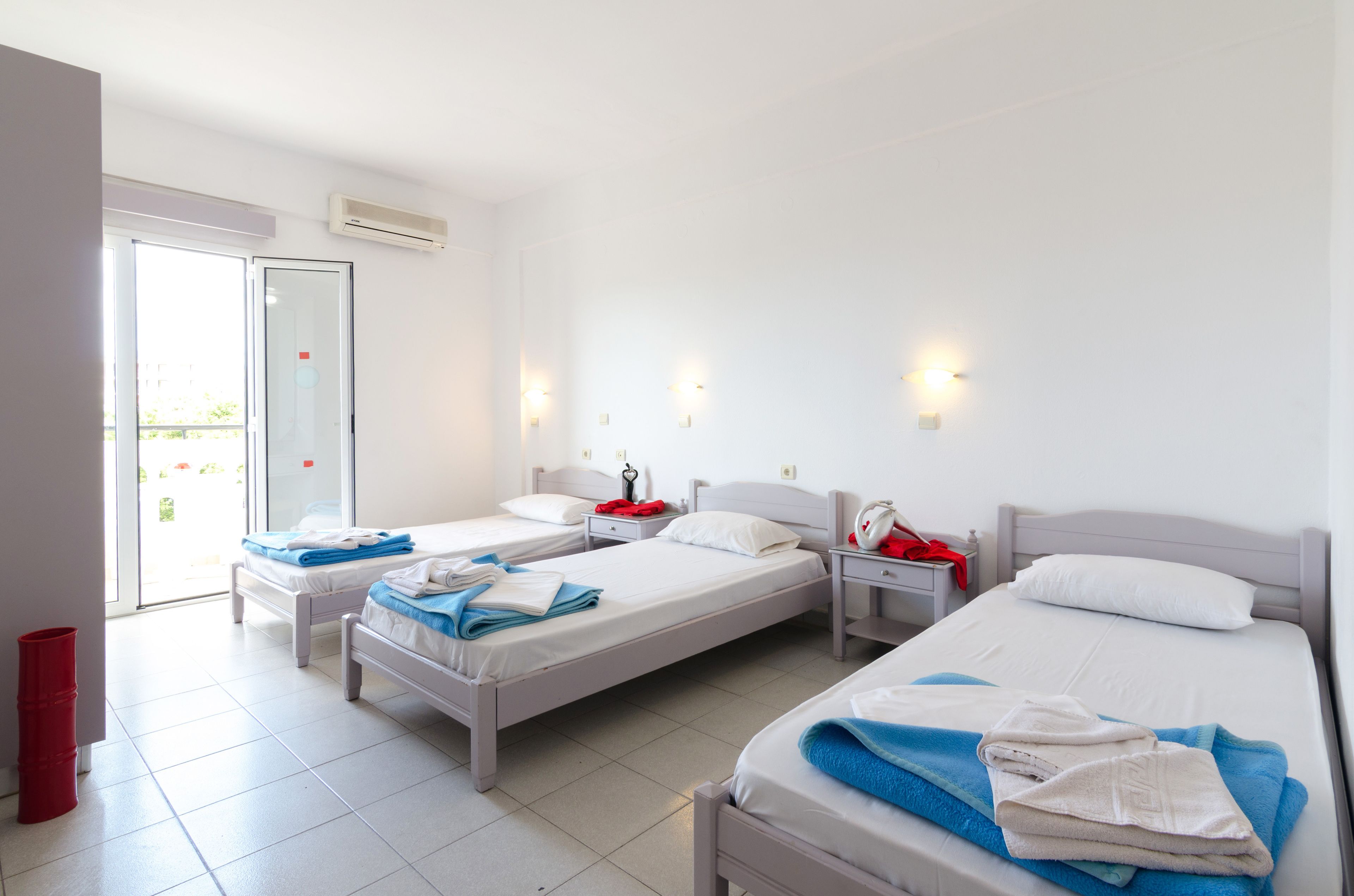 A hotel room with three single beds, a red trash bin, and a sliding glass door leading to a balcony.