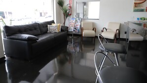 Un espacio de oficina moderno con un sofá de cuero negro, un sillón blanco y una silla negra. Hay una planta, un extintor y un revistero.