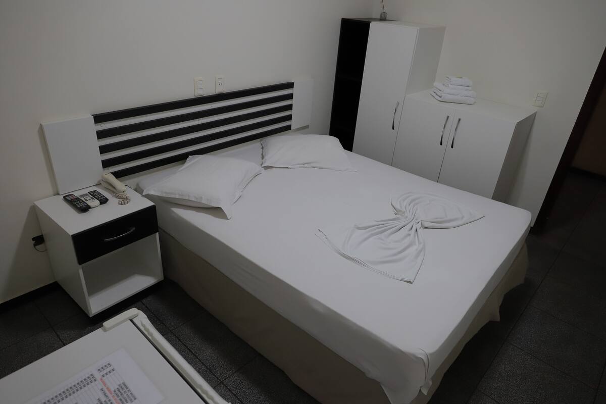 A neatly made bed with white linens, a bedside table with a phone and remote controls, a nightstand, and a wardrobe with towels on top.