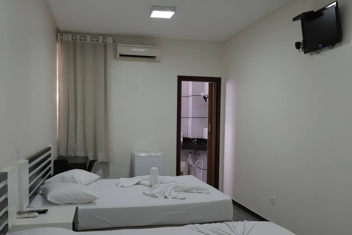 A hotel room with two beds, a flat-screen TV mounted on the wall, a desk with a chair, and a window with curtains.