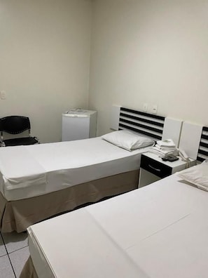 A hotel room with two beds, a desk, a chair, a small refrigerator, and a bedside table with a lamp. - Oft Neve's Hotel (Goiania)