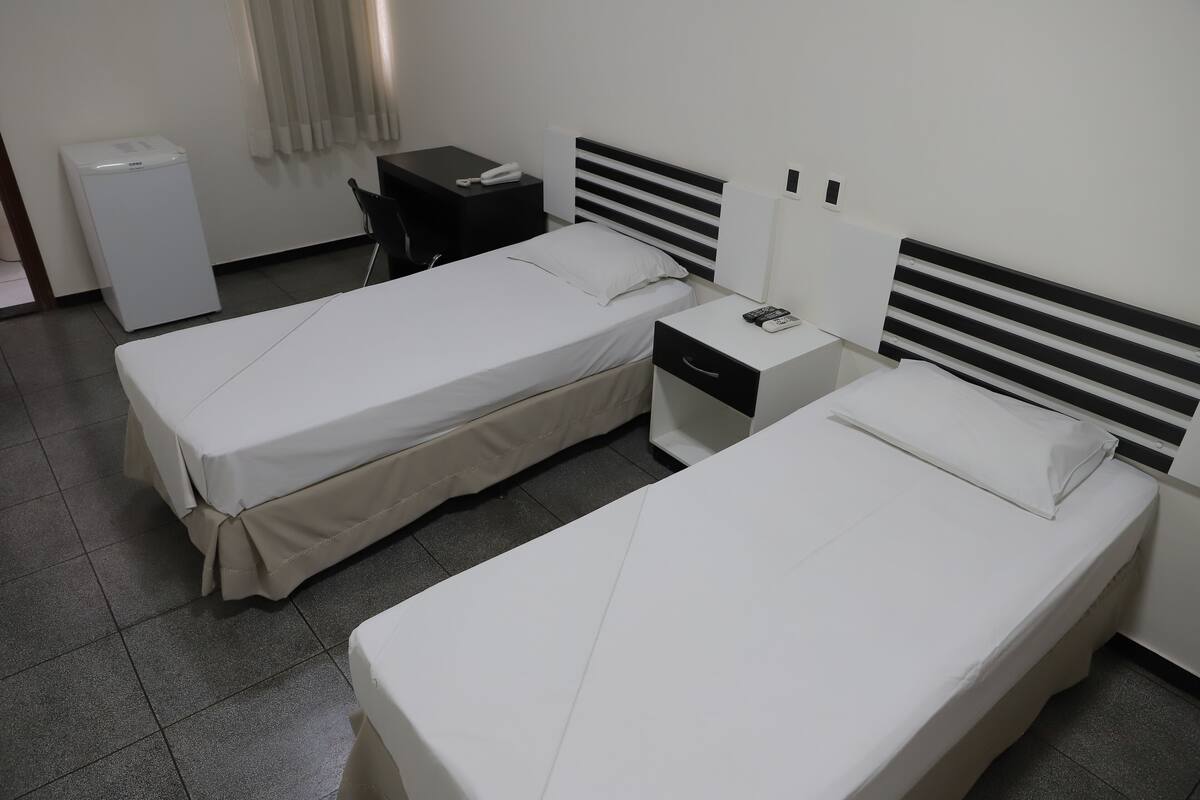 Two single beds with white linens, a small bedside table with a phone, a desk with a chair, and a white appliance.