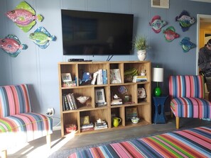 A living room with a TV, striped chairs, a bookshelf, and decorative fish on the wall. - Next 2 Paradise-203 2 Bedroom Condo by RedAwning (Supply)