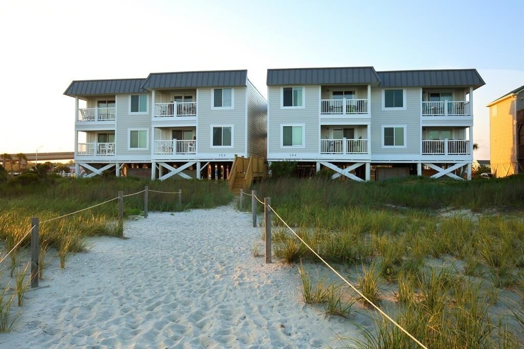 Un hôtel en bord de mer, avec des bâtiments à deux étages, un chemin de sable menant à l’entrée et une terrasse en bois.