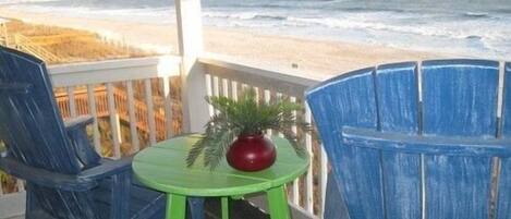Un balcón con vista a la playa, dos sillas azules, una mesa verde y un florero rojo.