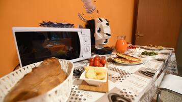 A breakfast table with a coffee machine, a bowl of tomatoes, a glass of orange juice, and various breakfast items.