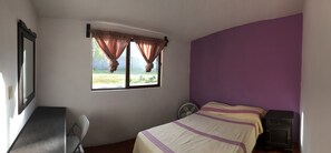 A bedroom with a bed, a desk, a chair, a window with curtains, and a fan.