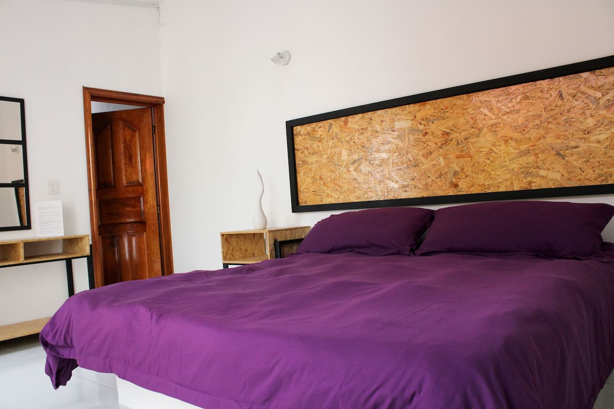 A bedroom with a large bed, a wooden door, a nightstand, and a wall-mounted artwork.