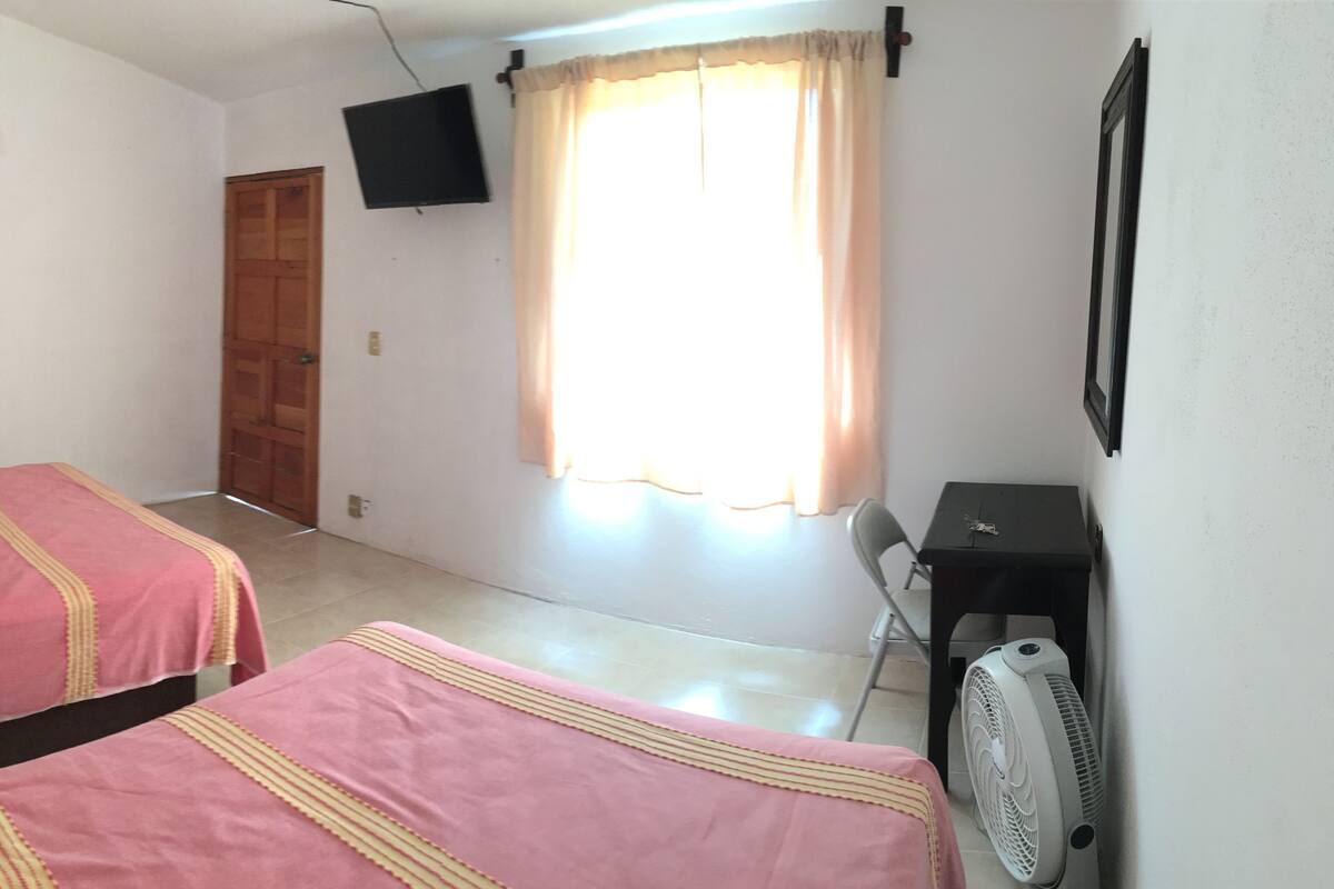 A room with two beds, a TV, a fan, and a window with curtains.