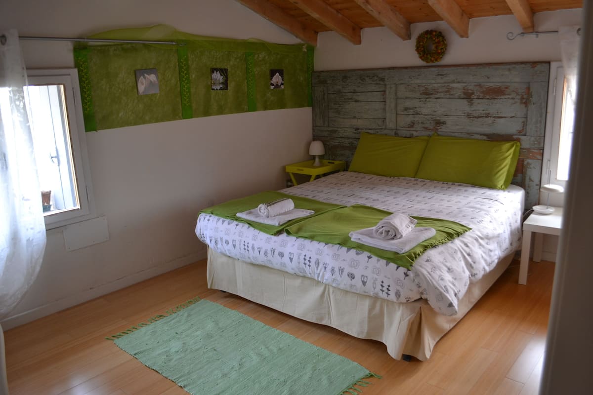 A bedroom with a wooden floor, a bed with green and white bedding, a small bedside table with a lamp, and a green wall with pictures.