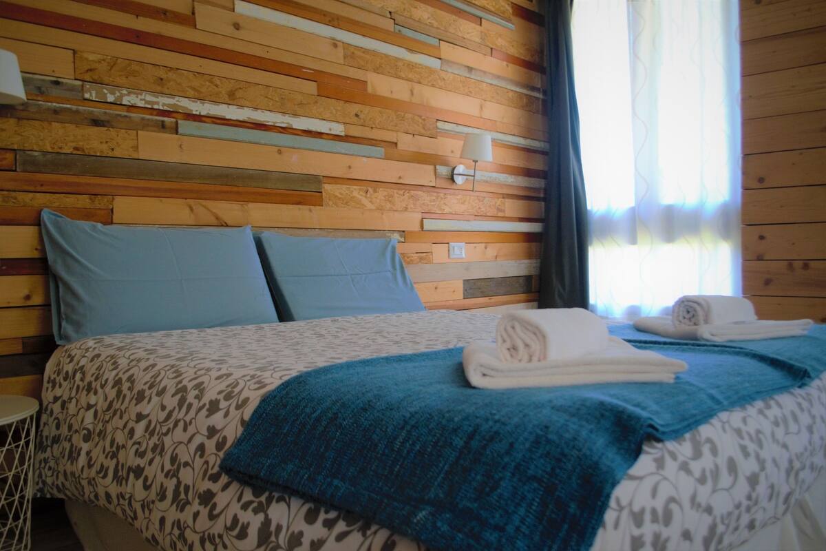 A room with a wooden slat wall, a bed with a patterned bedspread, two pillows, a blue throw blanket, and a rolled towel on the bed.