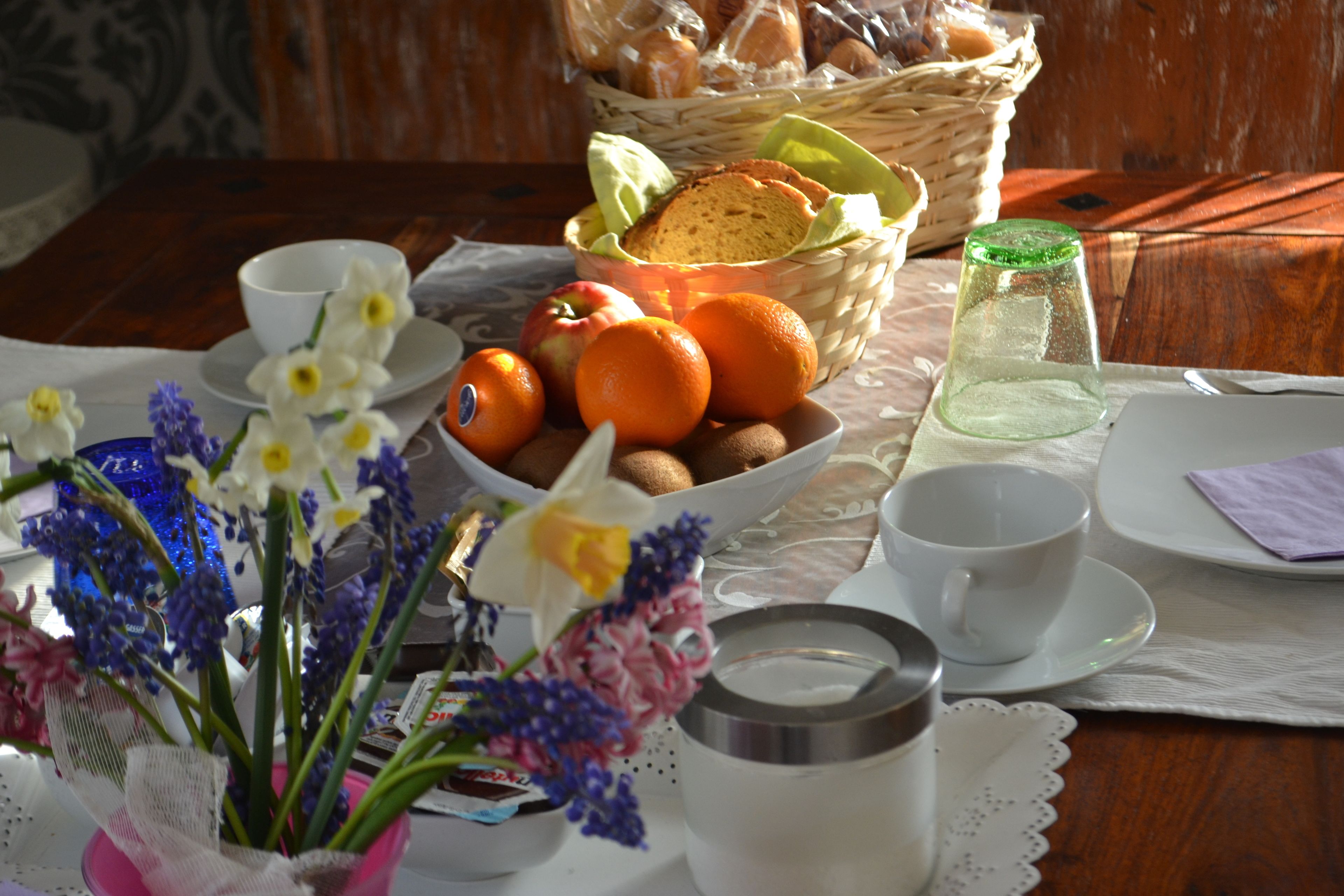 Un tavolo con una ciotola di arance, un cesto di pane, un bicchiere d'acqua e un vaso di fiori.