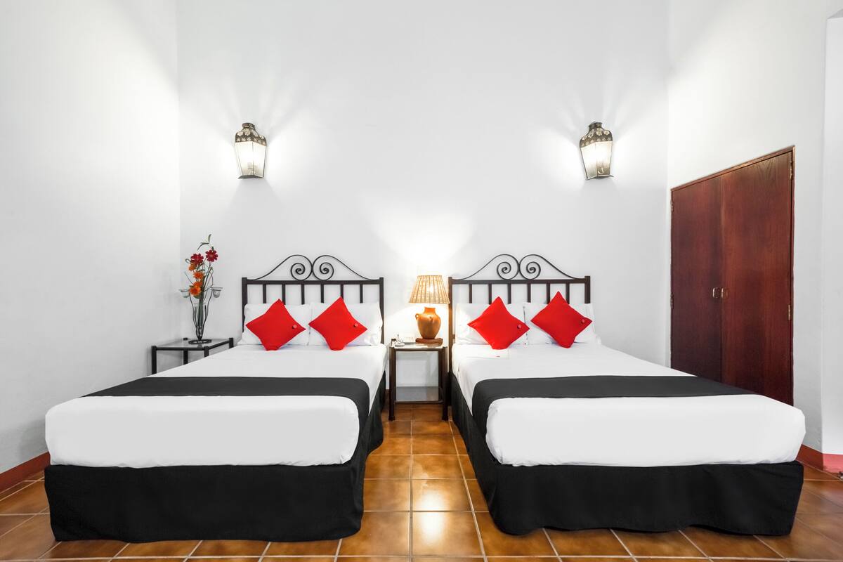 Una habitación de hotel con dos camas, ropa de cama blanca, almohadas rojas, una mesita de noche con lámpara y un jarrón con flores.