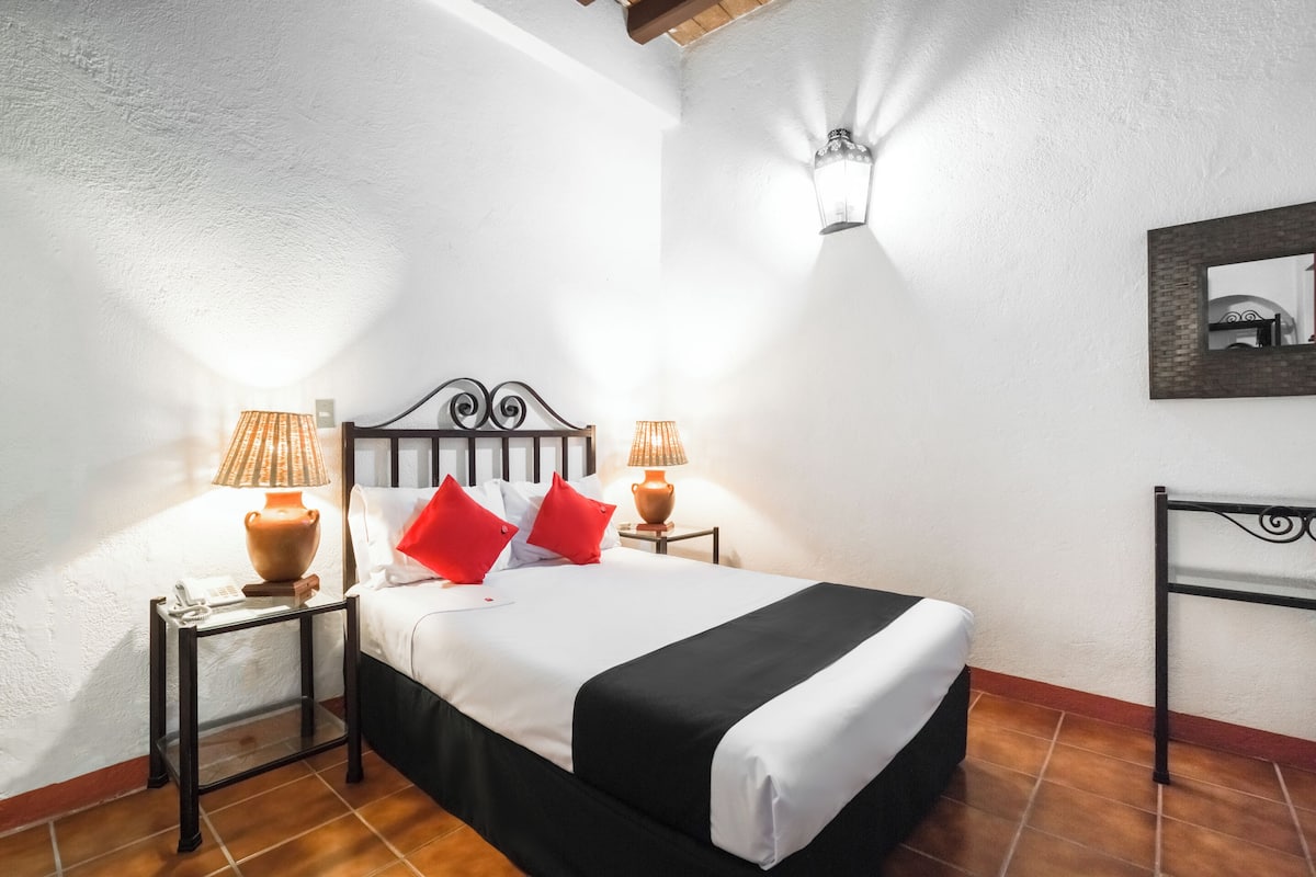 Una cama bien tendida con una colcha blanco y negro, dos almohadas rojas y dos lámparas de noche sobre mesitas auxiliares a juego.