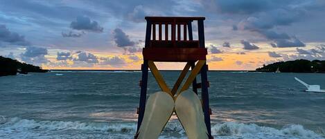 Una silla de playa con toldo blanco en la orilla, mirando al océano con olas y un atardecer de fondo.