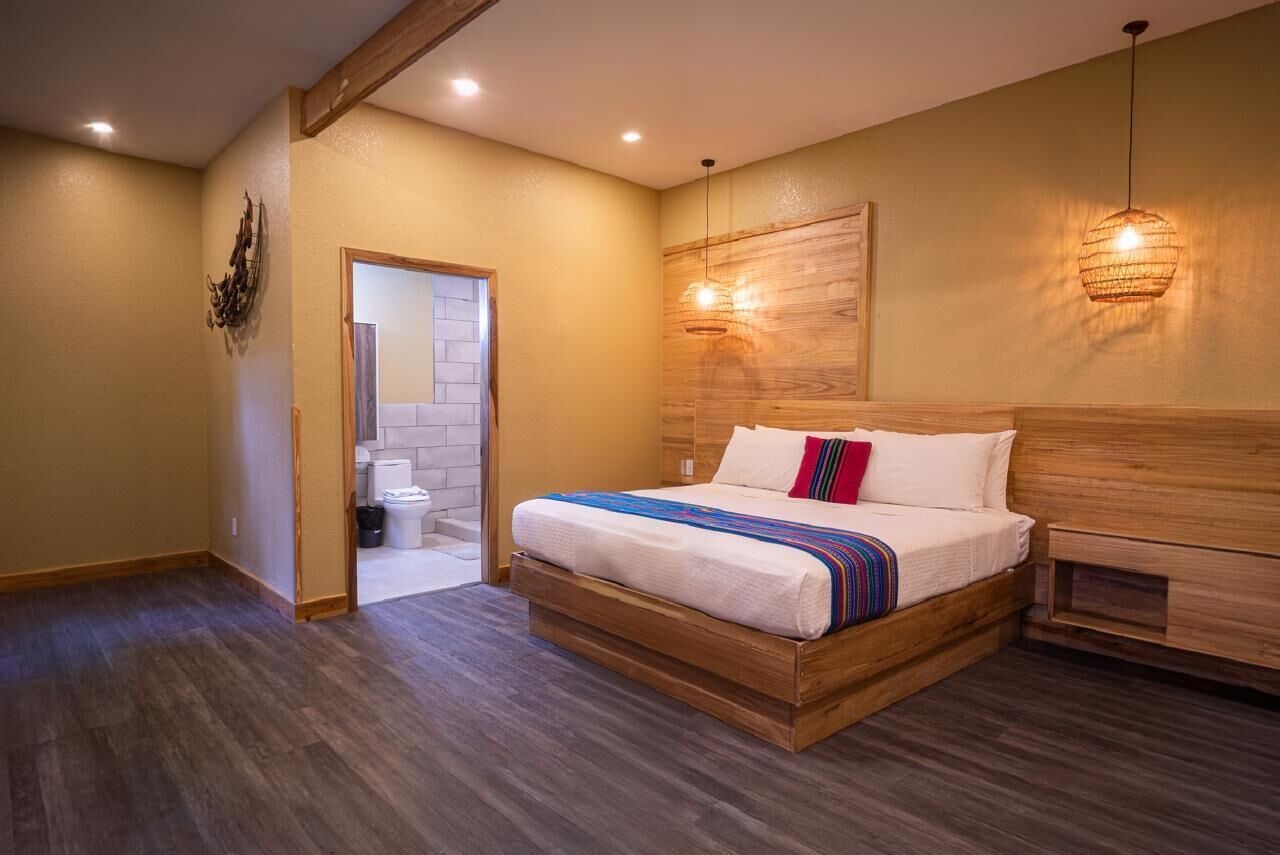 Un dormitorio con cama de madera, cabecera de madera, piso de madera, una lámpara fijada en la pared y una puerta que da a un baño.