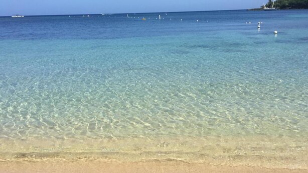A clear beach with shallow water, a sandy shore, and distant boats.