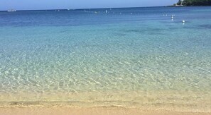 A clear beach with shallow water, a sandy shore, and distant boats.