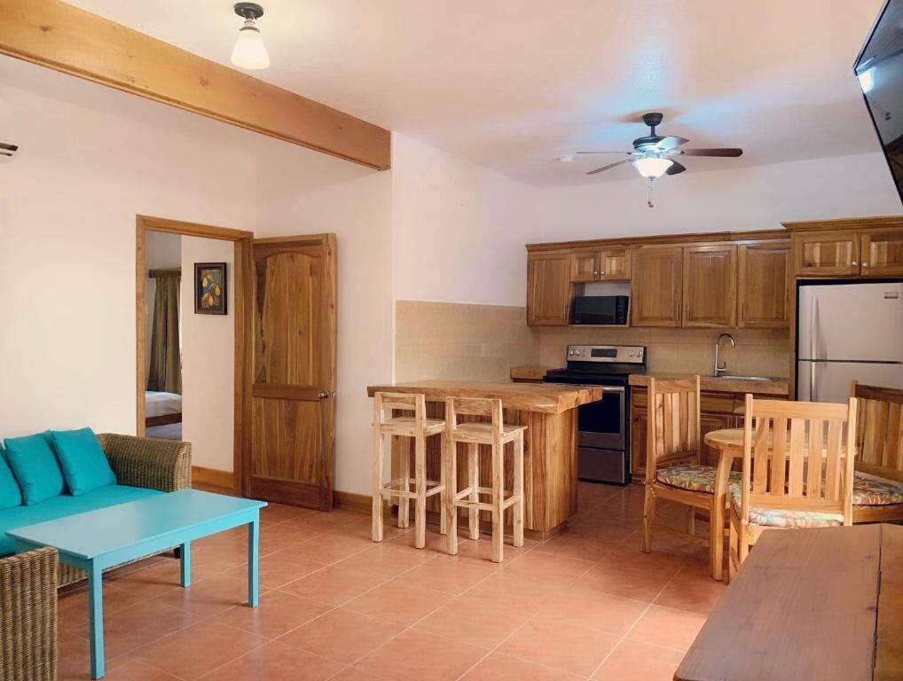 Cocina compacta con armarios de madera, microondas y refrigerador. Hay un comedor con mesa azul y sillas de madera.