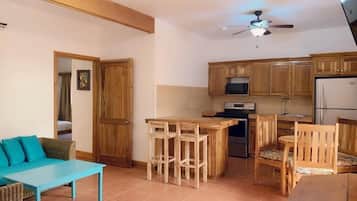 Cocina compacta con armarios de madera, microondas y refrigerador. Hay un comedor con mesa azul y sillas de madera.