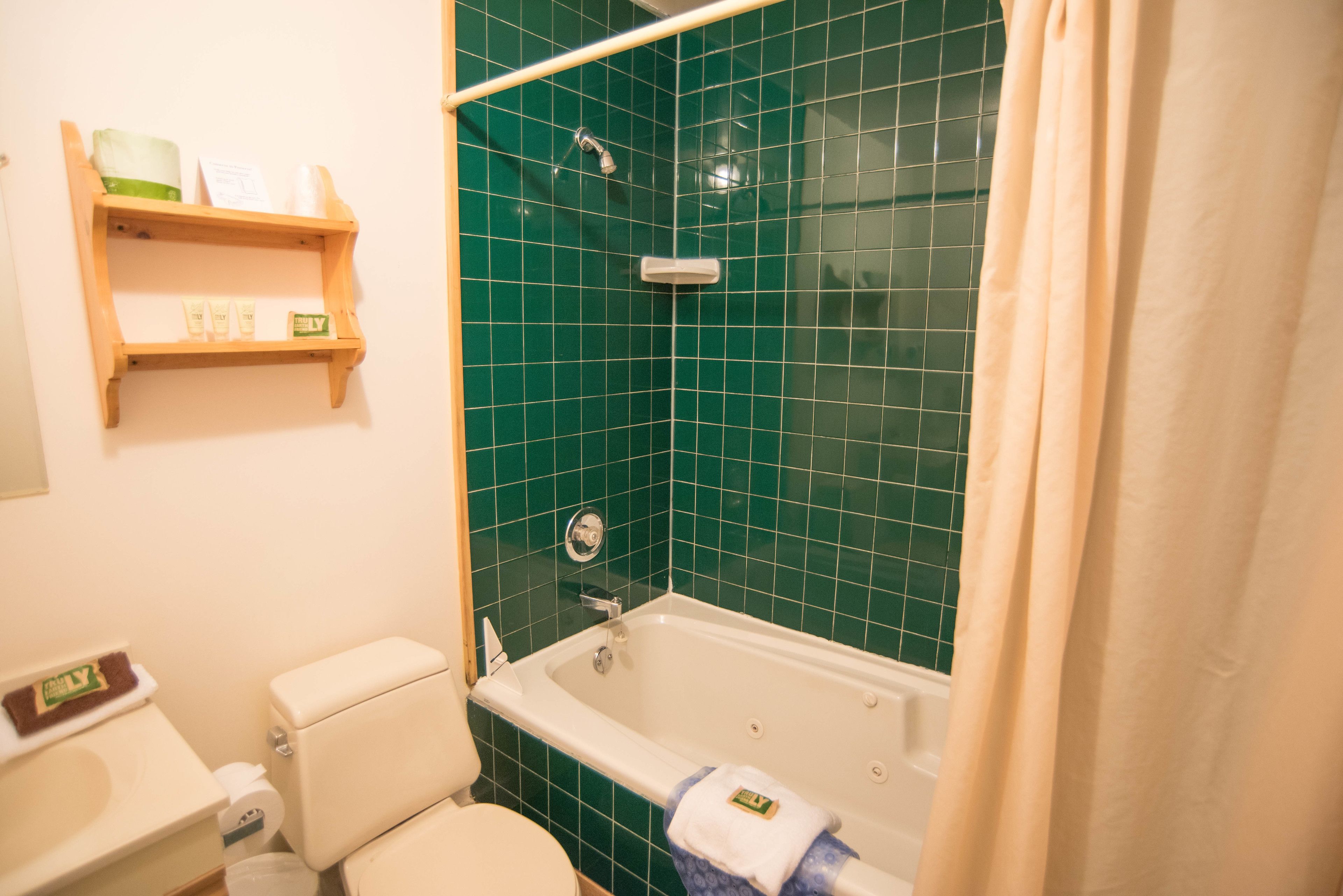 A bathroom with a bathtub, toilet, and green-tiled walls. There are shelves with toiletries and a towel hanging on the bathtub.