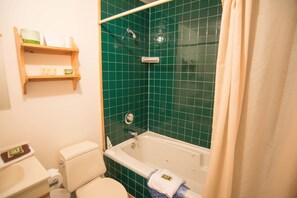 Une salle de bain avec une baignoire, des toilettes et des murs recouverts de carreaux verts. On y trouve des étagères avec des articles de toilette et une serviette suspendue à la baignoire.
