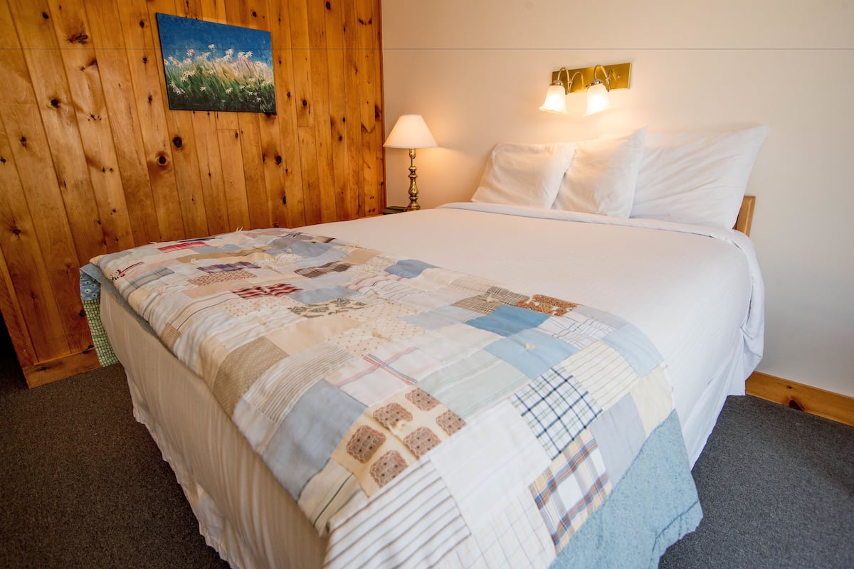 Cama bem arrumada com colcha, parede de madeira com pintura e luminária fixada na parede.