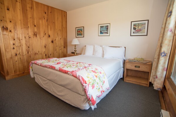A bedroom with a wooden panel wall, a bed with a floral bedspread, a nightstand with a lamp, and two framed pictures on the wall.