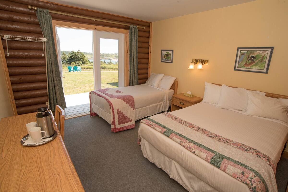 Quarto de hotel com duas camas, uma mesa de madeira com cafeteira e xícaras, e vista para o exterior por uma porta de correr de vidro.
